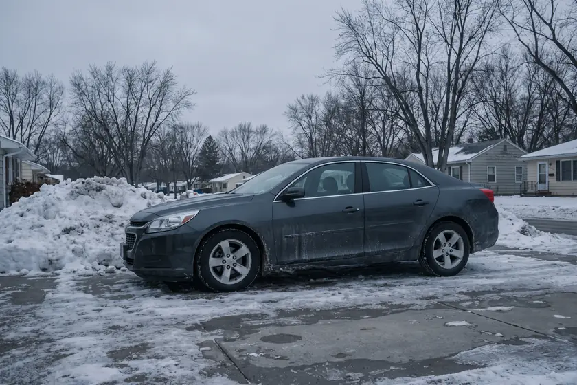 A grey sedan on a wet driveway beside a plowed snowbank